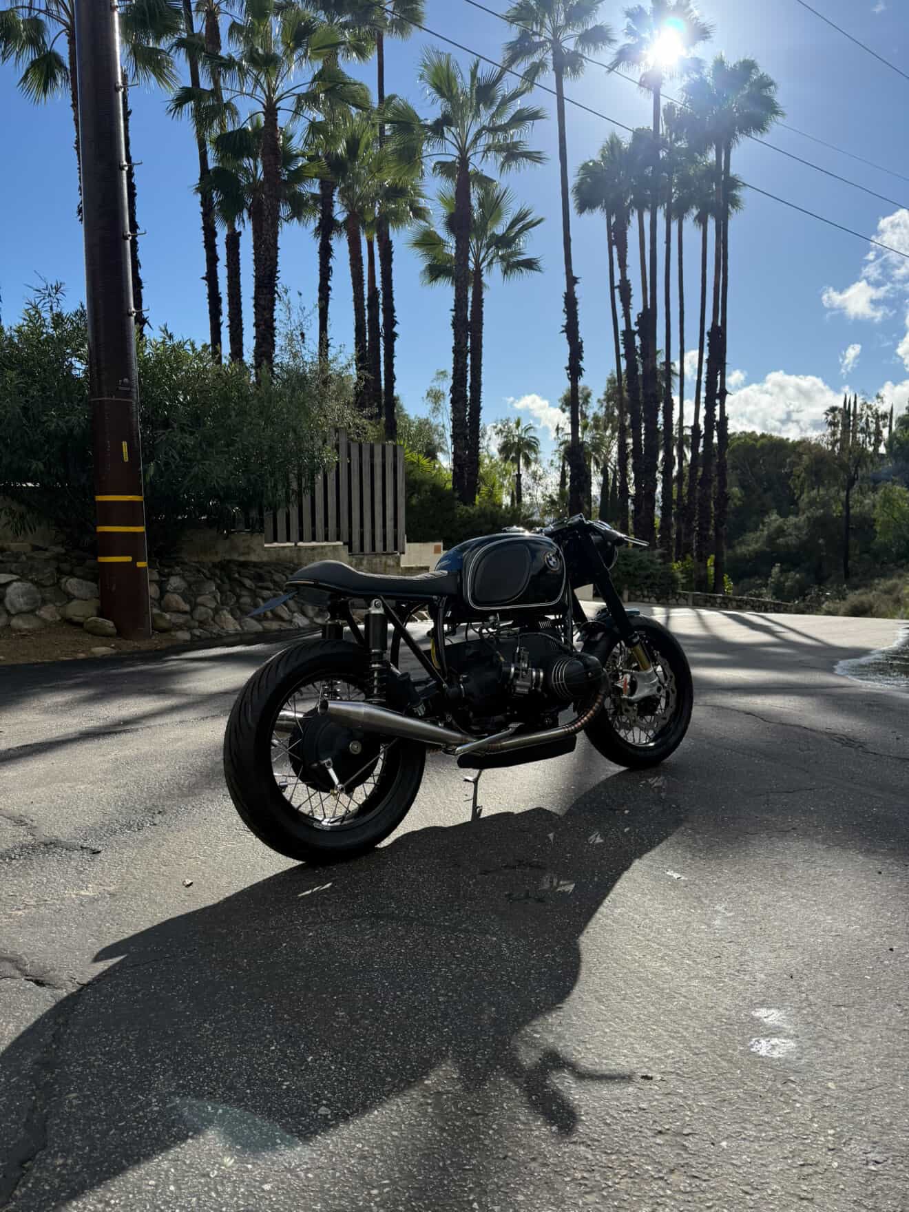 Rear-left view of the BMW airhead cafe racer showing the MAD Exhausts silencer, sunny palm-lined road