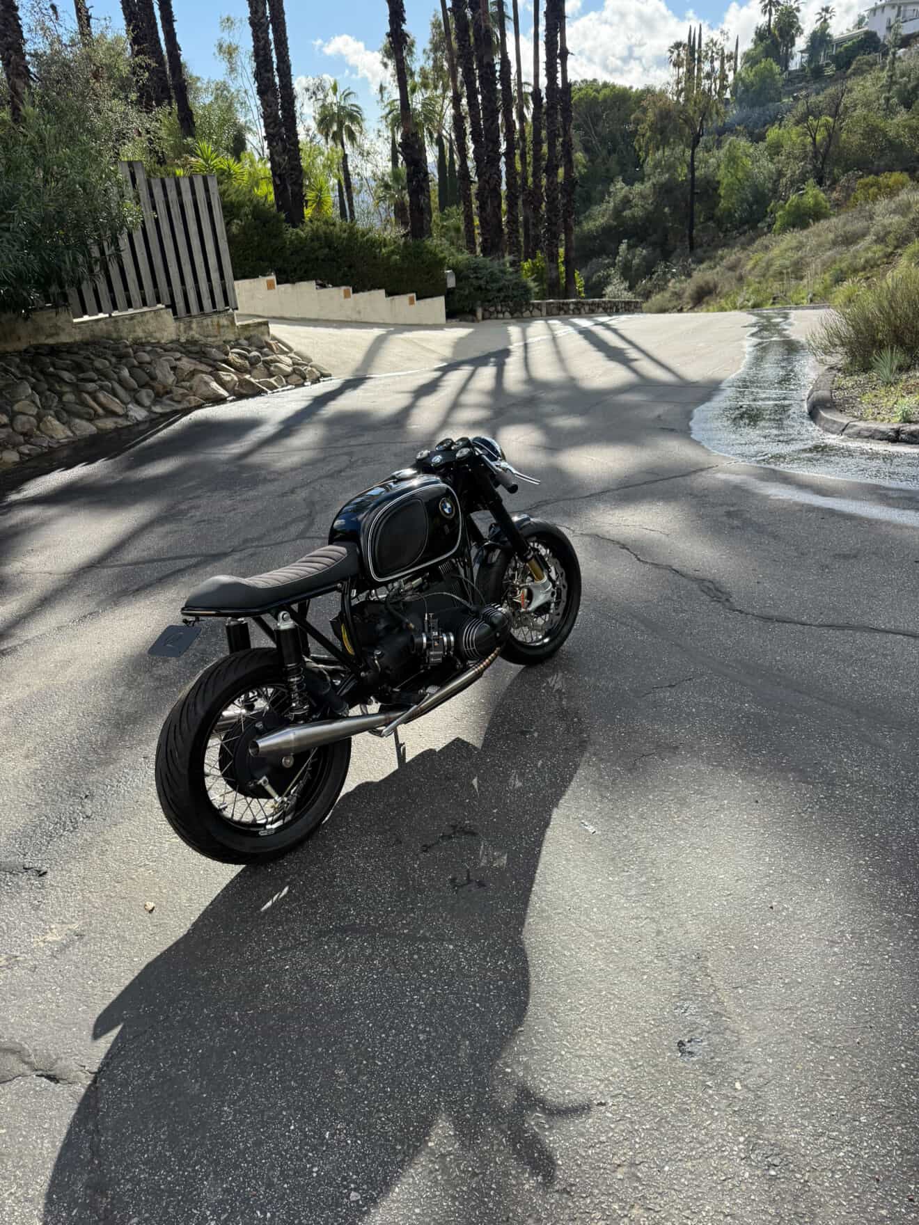 BMW airhead cafe racer with MAD Exhausts system on a hillside road, rear-left angle in dappled light